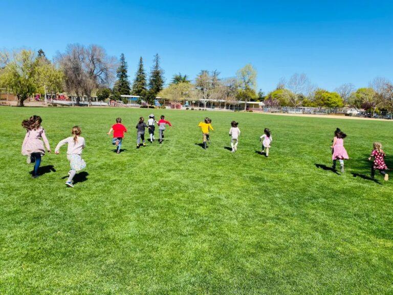 Children running across a wide grassy field on a sunny day.
